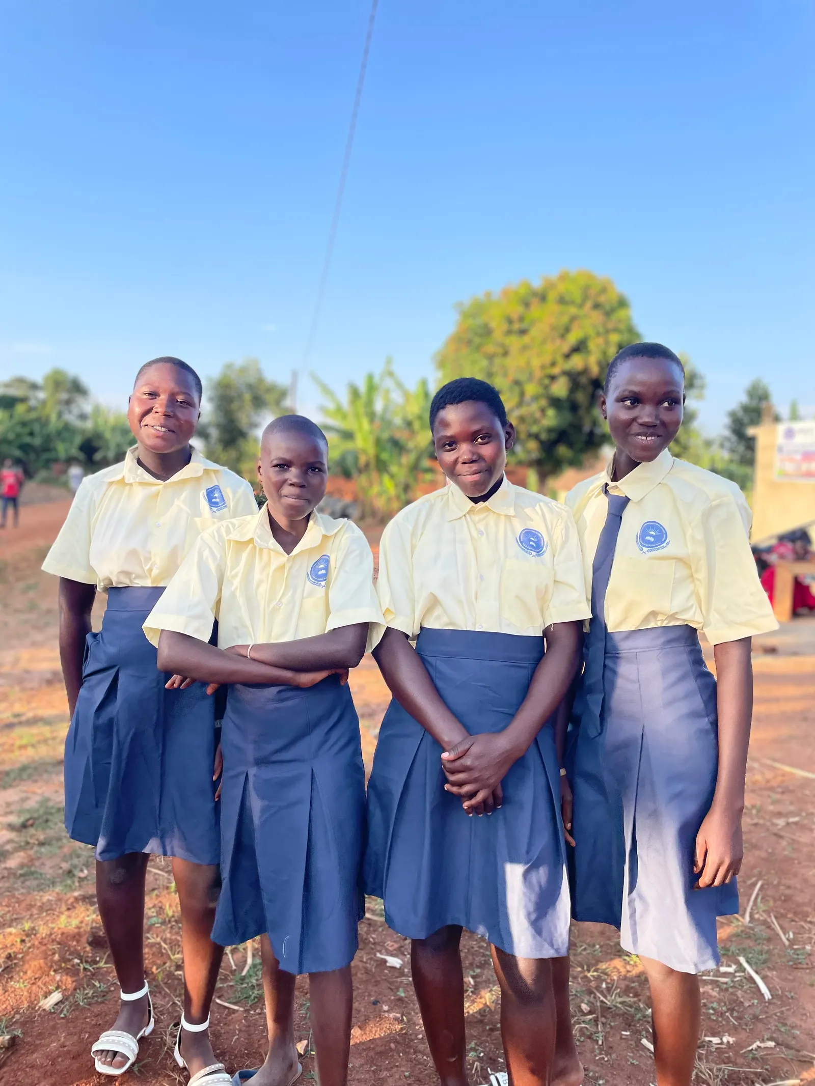 Four students in yellow shirts and navy skirts smiling in front of the school sign