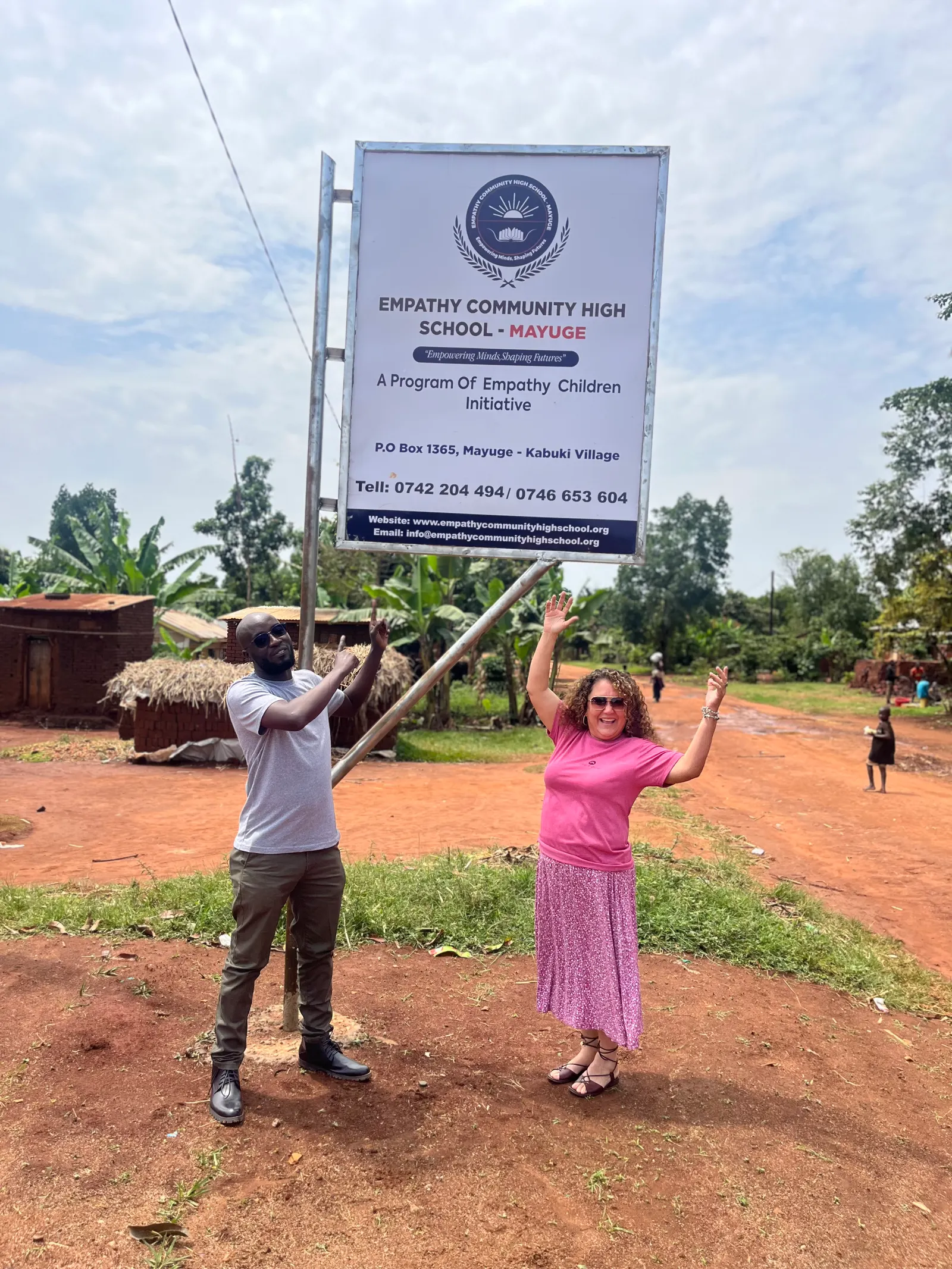 Two visitors stand beside the Empathy Community High School signboard on the road in Mayuge