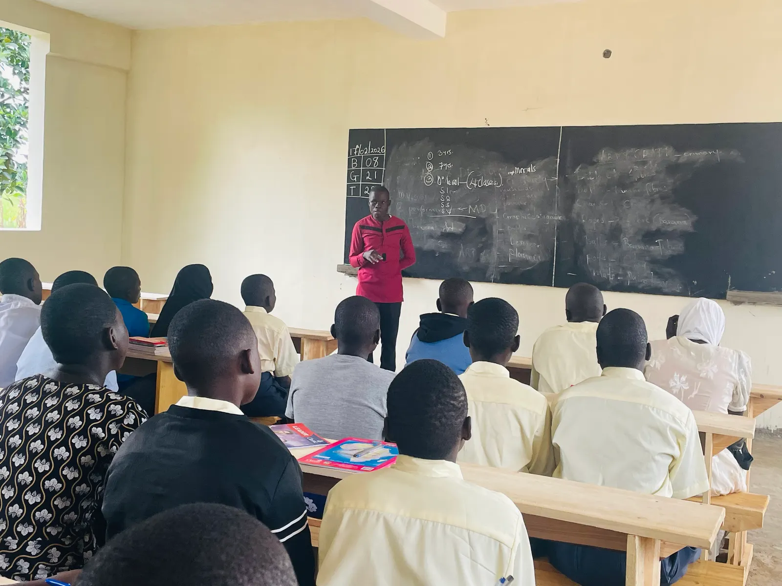 A teacher in a red top writes on the blackboard while students watch from their desks